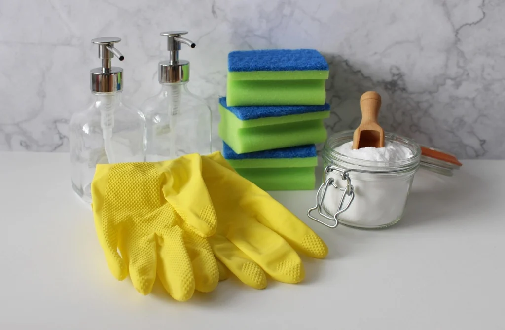 Yellow cleaning gloves, green and blue sponges, glass soap dispensers, and a jar of baking soda with a wooden scoop on a white surface.